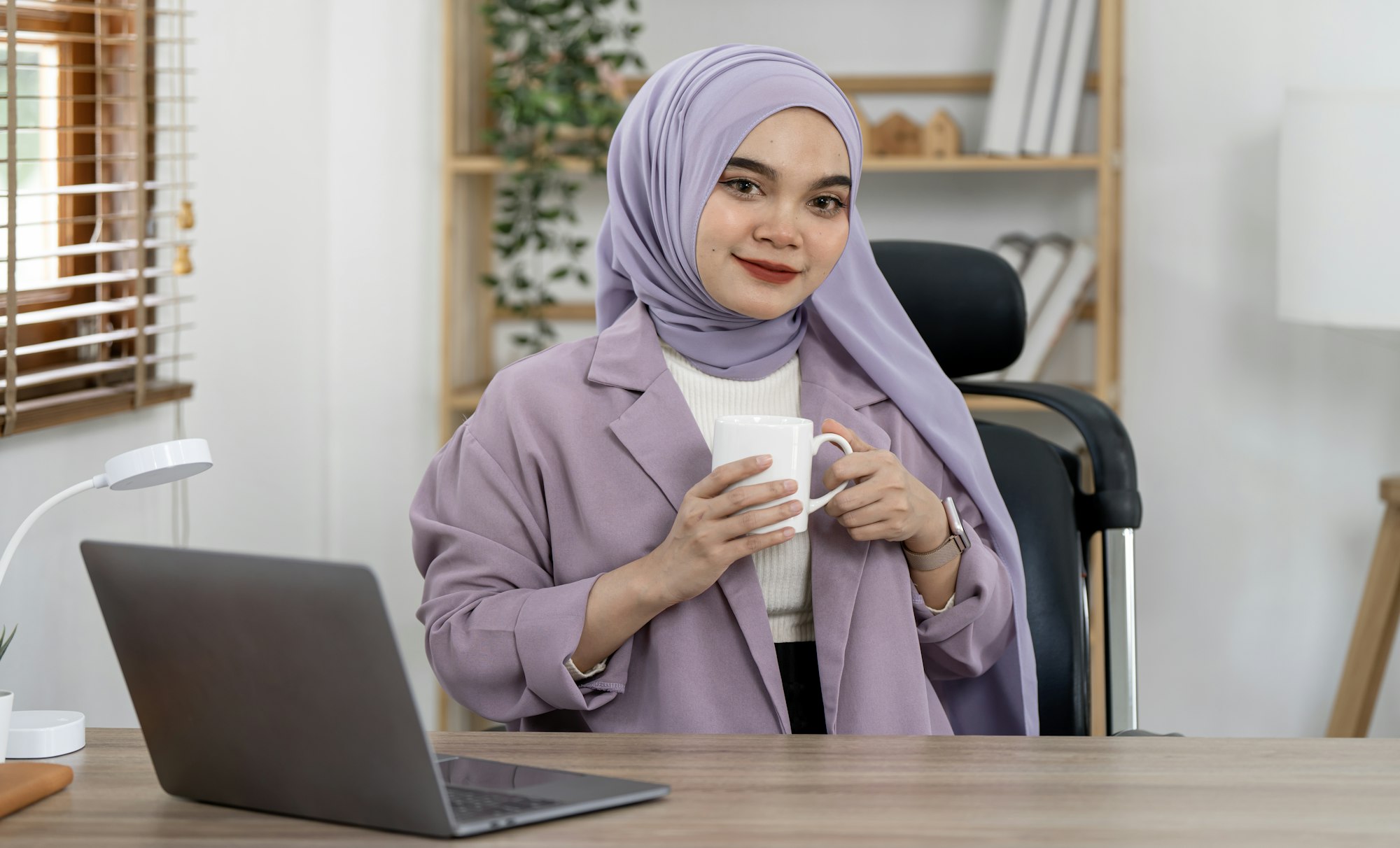 Smiling muslim businesswoman holding coffee cup sitting at the desk in home office space. Smiling muslim businesswoman holding coffee cup sitting at the desk in home office space.