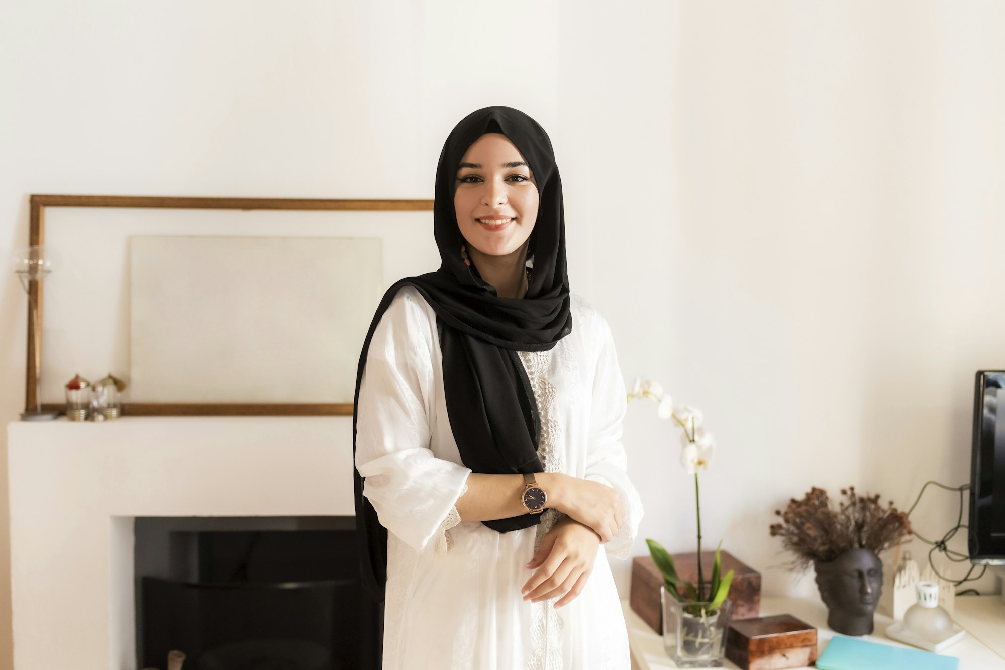 Smiling woman in a chic hijab standing in a cozy living room.